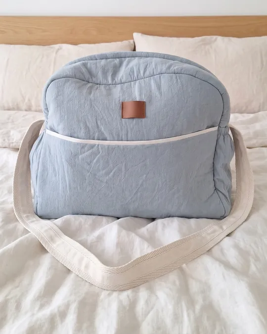 Bolso maternal acolchado celeste viejo de tusor de algodón, de frente, con bolsillo grande y correa cruda, sobre una cama.