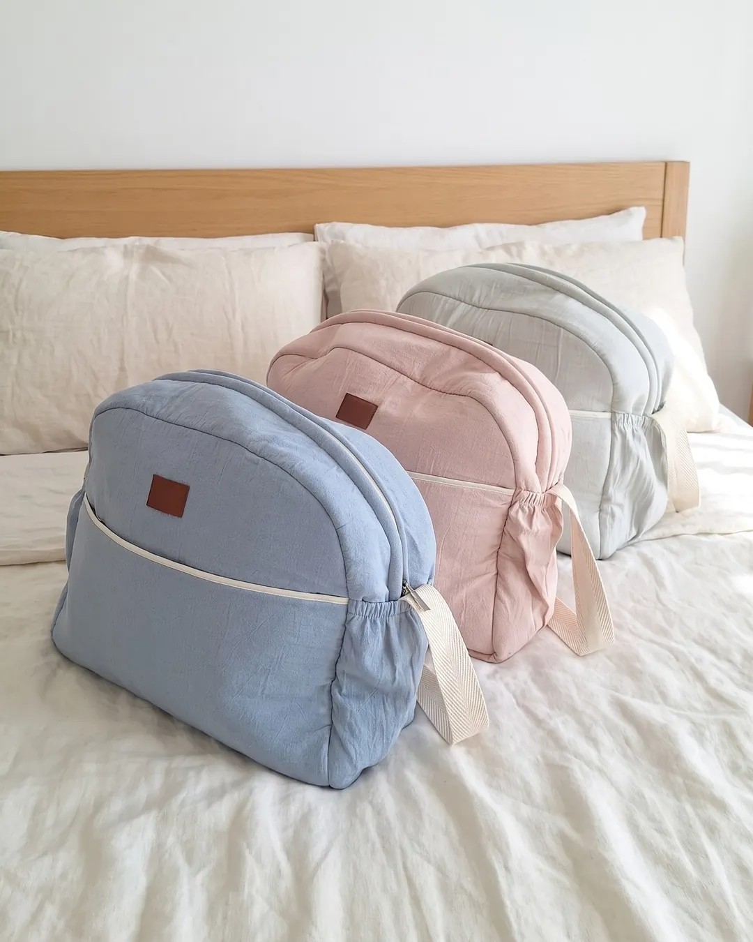 Tres bolsos maternales acolchados en celeste viejo, rosa viejo y gris perla, parados en fila sobre una cama.
